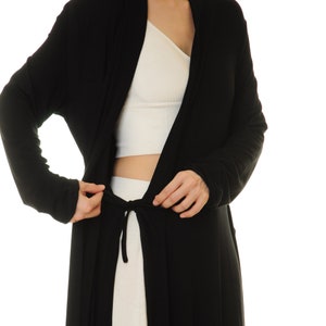 K&ouml;nnte beinhalten: Ein schwarzer Cardigan mit langen &Auml;rmeln und einem Bindeg&uuml;rtel. Der Cardigan ist vorne offen und zeigt darunter ein wei&szlig;es Crop-Top.