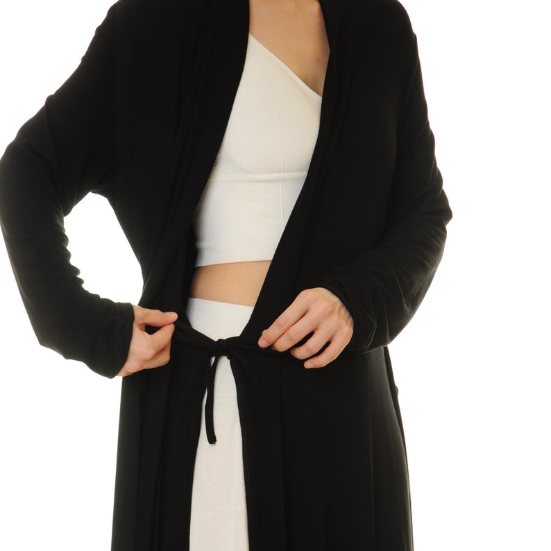 K&ouml;nnte beinhalten: Ein schwarzer Cardigan mit langen &Auml;rmeln und einem Bindeg&uuml;rtel. Der Cardigan ist vorne offen und zeigt darunter ein wei&szlig;es Crop-Top.