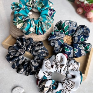 Puede incluir: Cuatro scrunchies estampados en turquesa, azul marino, negro y blanco. Los scrunchies están hechos de tela con un diseño de paisley.