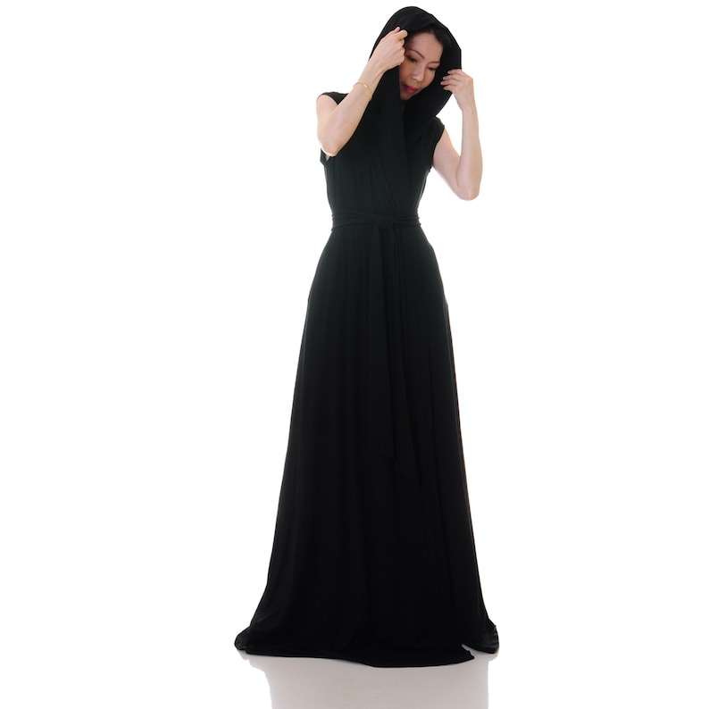 May include: A woman wearing a long, black maxi dress with a hood. The dress has a wrap style and a belt at the waist.