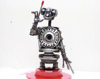 May include: A robot sculpture made of metal parts, including gears, bolts, and nuts. The robot is painted silver and has blue eyes. It is standing on a red base and has a red rod extending from its head.