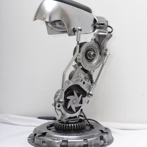 Könnte beinhalten: Eine einzigartige Schreibtischlampe aus recycelten Autoteilen. Die Lampe hat einen silberfarbenen Metallschirm mit schwarzem Oberteil, der von einer komplexen Anordnung aus Zahnrädern, Lagern und anderen mechanischen Komponenten getragen wird. Der Sockel ist eine kreisförmige Metallplatte.