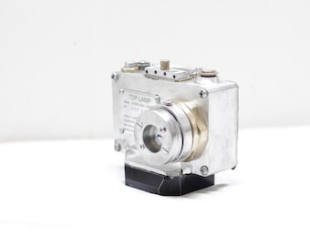 May include: A silver-colored industrial device with a cylindrical lens and a black base. The device has a rectangular body with screws and a label that reads "TOP LAMP". The lens is surrounded by a brass-colored ring.