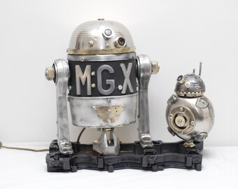May include: A pair of steampunk-style robot sculptures. The larger robot has the letters "M.G.X." on its body. The robots are made of metal and sit on a black base. The robots have a silver and gold color scheme.