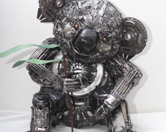 May include: A metal sculpture of a koala bear, crafted from various metal parts. The koala is depicted holding a branch with green leaves. The sculpture is primarily silver and gray, with details like the eyes and nose.