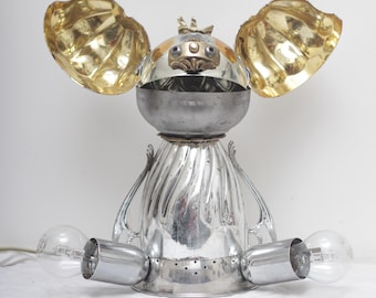 May include: A whimsical, handmade lamp sculpture. The lamp features a silver body with two light bulbs and gold-colored accents. The design resembles a creature with large ears and a crown.