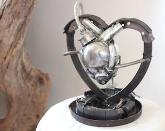 May include: A metal sculpture of a heart made from recycled car parts. The heart is silver and the base is black.