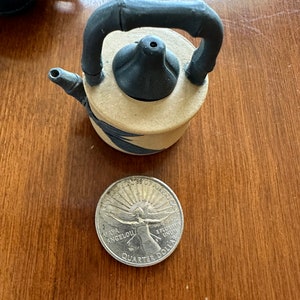 May include: A small ceramic teapot with a black handle and lid, and a tan body with blue leaf accents. A Maya Angelou quarter is next to the teapot.