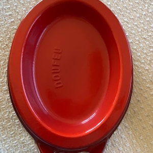 May include: A red oval-shaped Doufeu Dutch oven with two handles. The interior is a glossy red, and the word "DOUFEU" is embossed on the interior base. This cookware is ideal for slow cooking and braising.