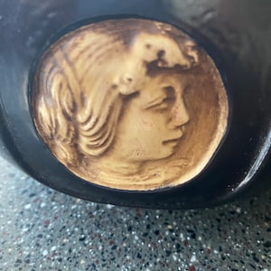 May include: A black ceramic bowl with a raised, cream-colored cameo of a woman's face. The woman's hair is styled in a wave and she is looking to the right.