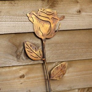May include: A metal rose with a gold finish, the rose is cut out of metal and has a rustic look. The rose is on a wooden background.