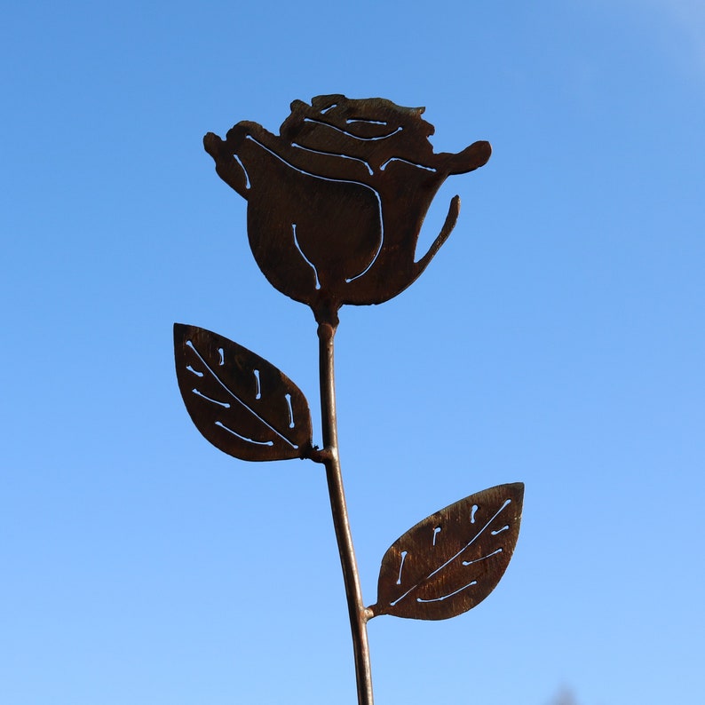 May include: A metal rose silhouette with two leaves, painted in a rusty brown color, against a blue sky background.