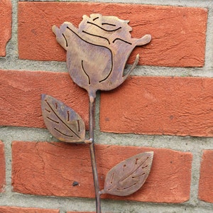 May include: A metal rose sculpture with a brown patina. The rose has a single stem with two leaves. The sculpture is mounted on a brick wall.