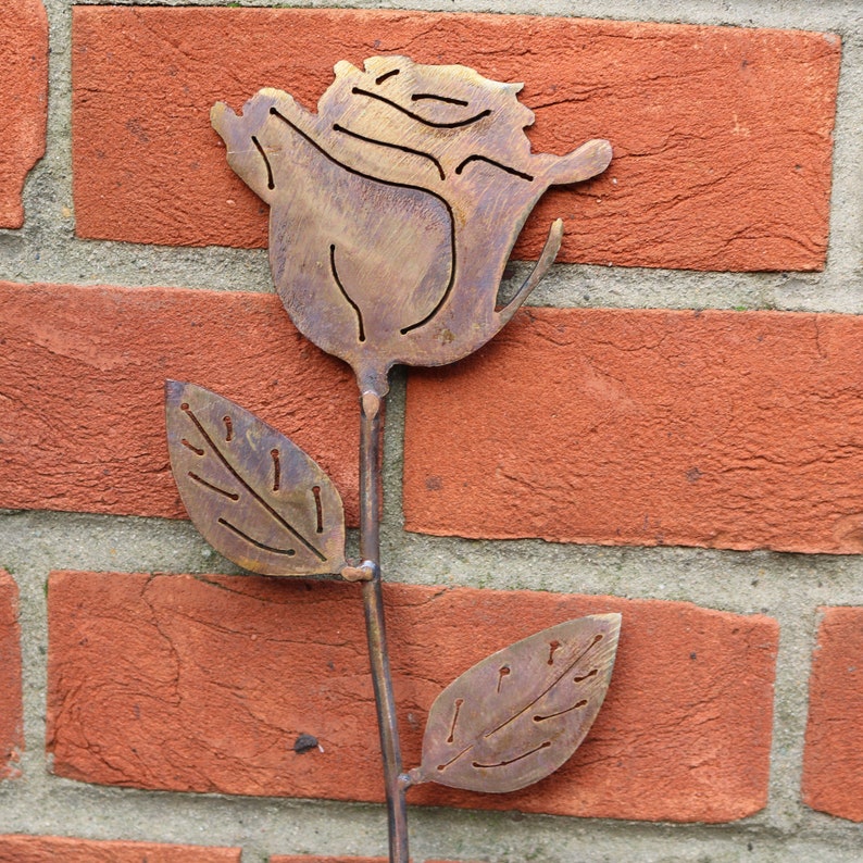 May include: A metal rose sculpture with a brown patina. The rose has a single stem with two leaves. The sculpture is mounted on a brick wall.
