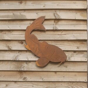 May include: A rusty metal fish sculpture mounted on a wooden wall. The fish is facing to the right and has a curved tail.