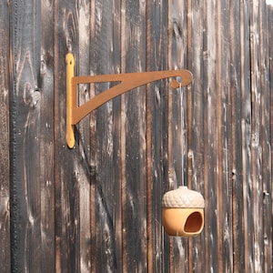 May include: A wooden hanging bracket with an acorn-shaped bird feeder. The bracket is painted in shades of yellow and brown, and is mounted on a weathered wooden fence. The bird feeder is tan and has a small opening.