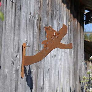 May include: A rusty metal frog sculpture mounted on a wooden wall. The frog is perched on a branch with its legs outstretched. The sculpture is a whimsical and decorative addition to any outdoor space.