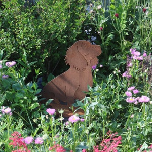 May include: A rusty metal silhouette of a dog sitting in a garden with pink flowers. The dog is facing to the right and has its head tilted up.