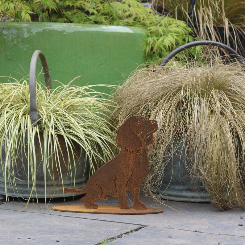 May include: A rusty metal silhouette of a dog sitting on a base. The dog is brown and has a long tail. The silhouette is in front of two rusty metal buckets with plants growing out of them.