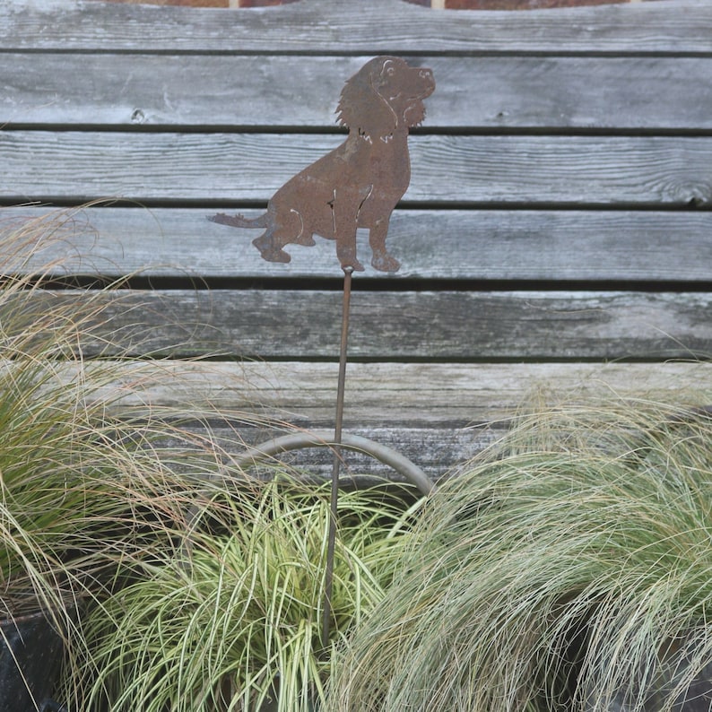 Op de afbeelding: Een metalen silhouet van een bruine hond, staand op een metalen paal, voor een houten achtergrond.