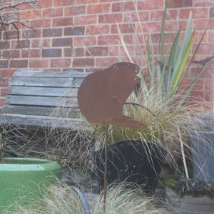 May include: A rusty metal silhouette of a beaver sitting on a metal rod, surrounded by tall green grass and a wooden bench in the background.