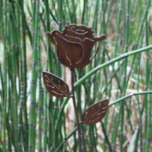 May include: A rusty metal rose with two leaves, the rose is cut out and the leaves are cut out with holes, the rose is in front of a green background.