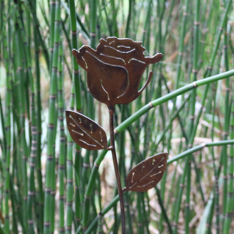 May include: A rusty metal rose with two leaves, the rose is cut out and the leaves are cut out with holes, the rose is in front of a green background.