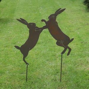 May include: Two metal silhouettes of rabbits in a fighting pose, mounted on stakes and placed in a grassy lawn.