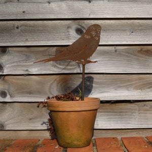 May include: A rusty metal bird sculpture in the shape of a parakeet perched on a metal stake. The sculpture is placed in a terracotta flower pot with dried leaves and twigs.