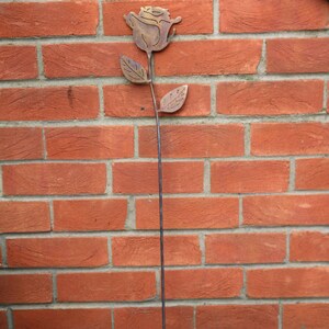 May include: A metal rose sculpture with a brown patina finish. The rose is attached to a long, thin metal stem. The sculpture is mounted on a brick wall.