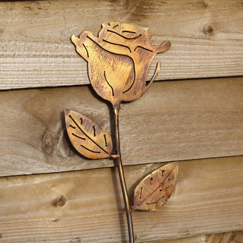 May include: A metal rose with a gold finish, the rose is cut out of metal and has a rustic look. The rose is on a wooden background.