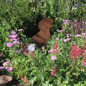 May include: A rusty metal silhouette of a dog standing in a garden with pink and purple flowers. The dog is facing to the right and has its head tilted up.