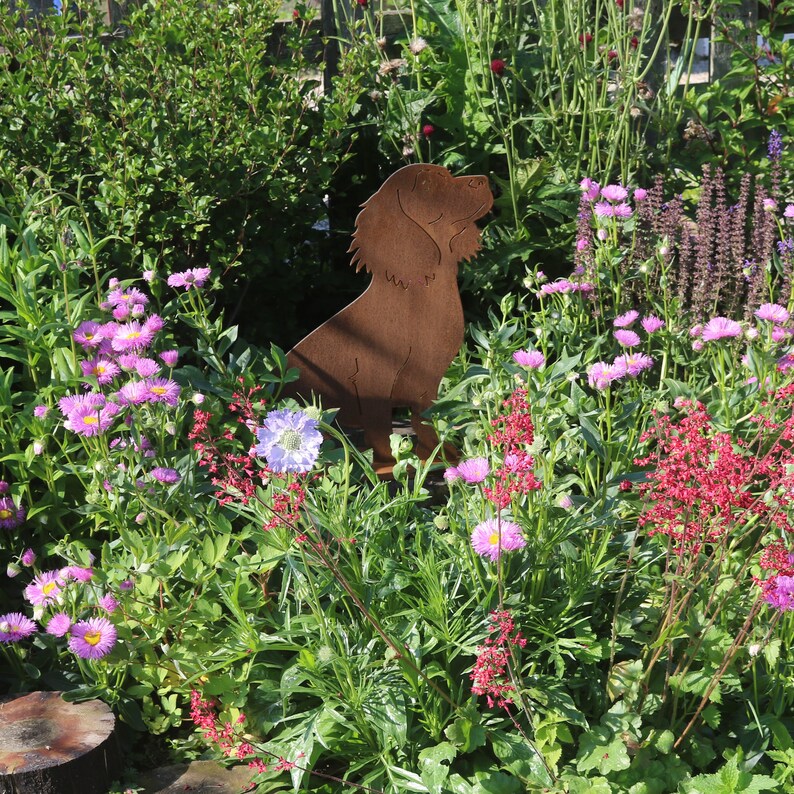 May include: A rusty metal silhouette of a dog standing in a garden with pink and purple flowers. The dog is facing to the right and has its head tilted up.