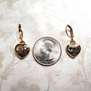 May include: A pair of gold-tone earrings with a heart-shaped design. Each heart features a clear resin filled with intricate gold and black gears.