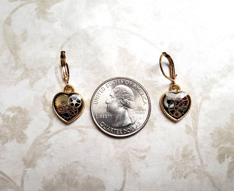 May include: A pair of gold-tone earrings with a heart-shaped design. Each heart features a clear resin filled with intricate gold and black gears.