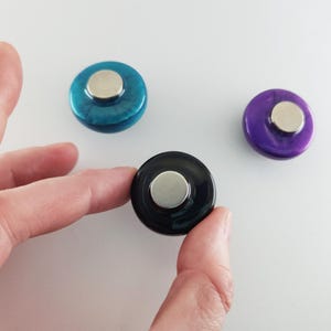 May include: Three circular magnets with smooth, glossy surfaces in teal, purple, and black. Each magnet features a raised, metallic silver-toned circular center piece on the front, providing a distinct contrast to the colored, rounded outer edges.