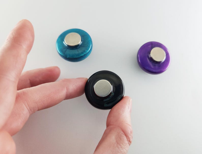 May include: Three circular magnets with smooth, glossy surfaces in teal, purple, and black. Each magnet features a raised, metallic silver-toned circular center piece on the front, providing a distinct contrast to the colored, rounded outer edges.
