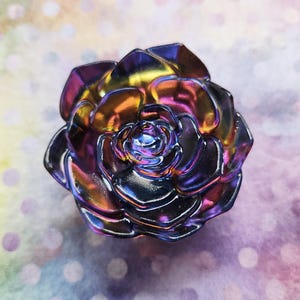 Könnte beinhalten: Ein mehrfarbiges, irisierendes, blumenförmiges Objekt mit metallischem Glanz. Die Blütenblätter zeigen einen Farbverlauf aus tiefem Violett, Blau und Goldgelb, angeordnet in einem geschichteten, kreisförmigen Muster, das einer blühenden Rose ähnelt.