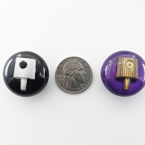 May include: Two circular buttons, one black and one purple, each featuring a spray paint nozzle cap embedded in the center. A United States quarter coin is placed between the buttons for scale, measuring approximately 0.955 inches in diameter.