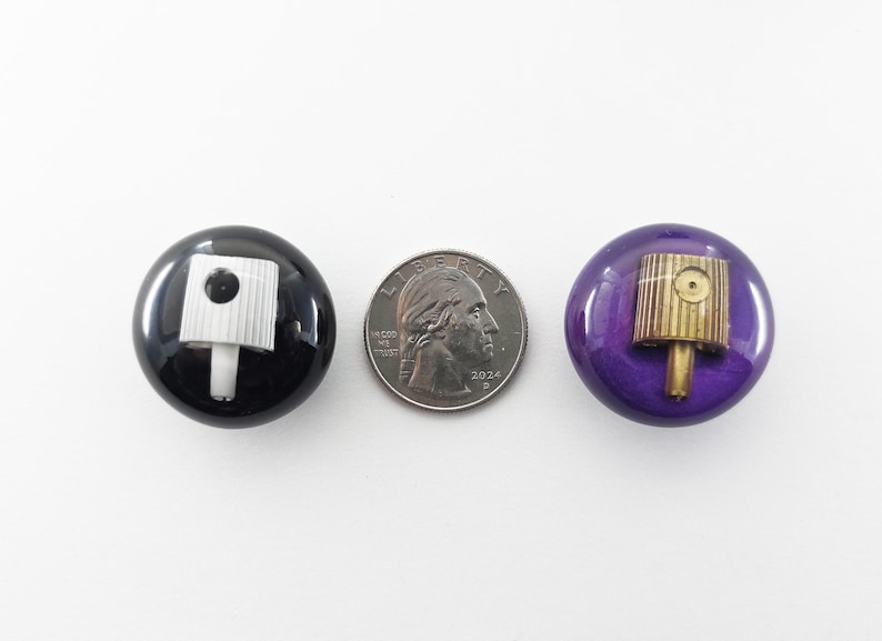 May include: Two circular buttons, one black and one purple, each featuring a spray paint nozzle cap embedded in the center. A United States quarter coin is placed between the buttons for scale, measuring approximately 0.955 inches in diameter.