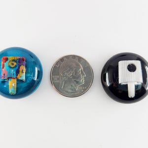 May include: Two circular spray paint can caps, one metallic blue with a multi-colored nozzle and one glossy black with a white nozzle, positioned on either side of a United States quarter coin for scale, measuring approximately 0.95 inches in diameter.