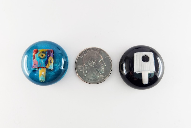 May include: Two circular spray paint can caps, one metallic blue with a multi-colored nozzle and one glossy black with a white nozzle, positioned on either side of a United States quarter coin for scale, measuring approximately 0.95 inches in diameter.