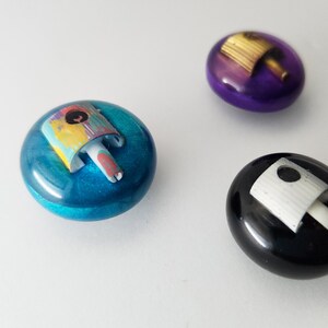 May include: Three circular, smooth, resin-like buttons or cabochons. One is metallic blue with a multi-colored spray paint nozzle cap, one is deep purple with a gold nozzle, and one is glossy black with a white spray paint nozzle cap.