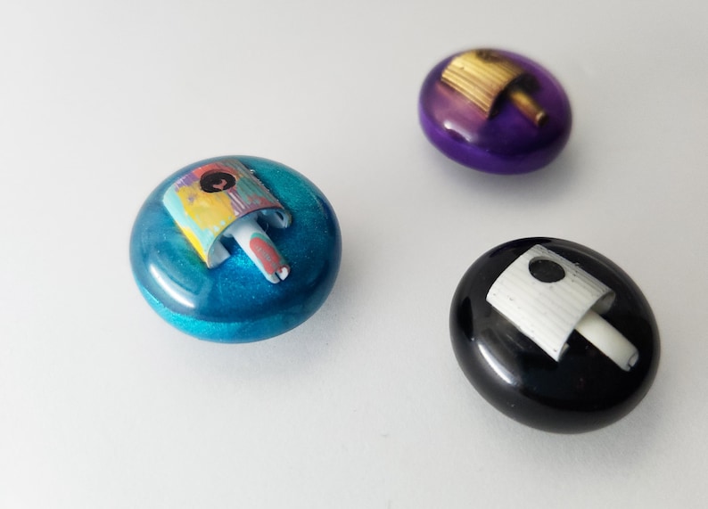 May include: Three circular, smooth, resin-like buttons or cabochons. One is metallic blue with a multi-colored spray paint nozzle cap, one is deep purple with a gold nozzle, and one is glossy black with a white spray paint nozzle cap.