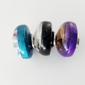 May include: Three cylindrical glass beads arranged horizontally. The left bead is teal with white and red accents. The center bead is black with white vertical stripes. The right bead is purple with gold-toned metallic vertical line details.