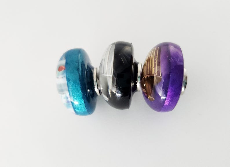 May include: Three cylindrical glass beads arranged horizontally. The left bead is teal with white and red accents. The center bead is black with white vertical stripes. The right bead is purple with gold-toned metallic vertical line details.