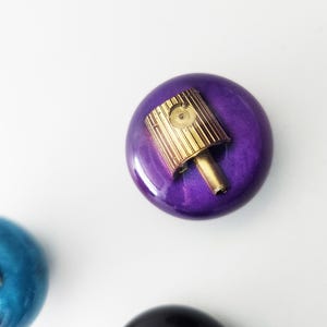 May include: A round, glossy purple object with a metallic gold spray paint nozzle cap attached to the top center. A second, partially visible blue object with a similar nozzle cap is positioned in the lower left corner of the frame.