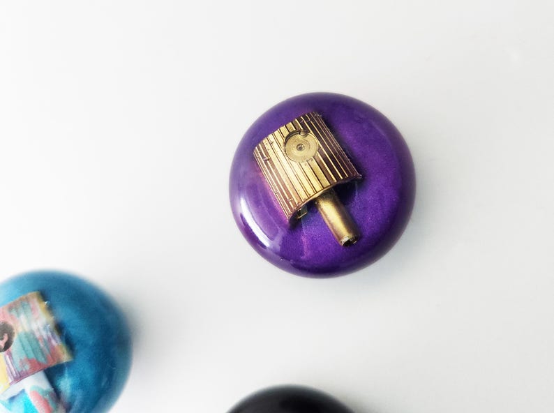 May include: A round, glossy purple object with a metallic gold spray paint nozzle cap attached to the top center. A second, partially visible blue object with a similar nozzle cap is positioned in the lower left corner of the frame.