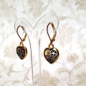 May include: A pair of gold-tone heart-shaped earrings with a black background and intricate gold and silver gears inside. The earrings have lever back closures.
