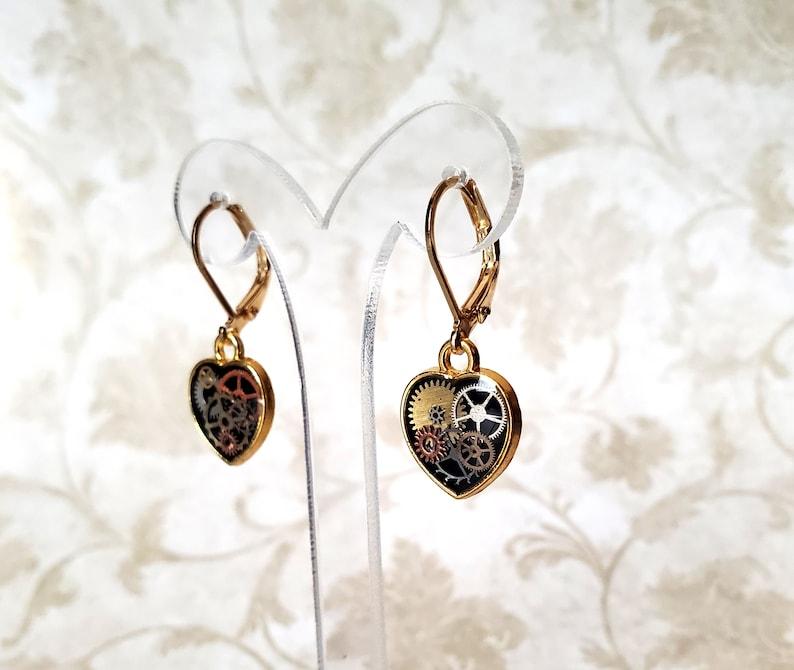 May include: A pair of gold-tone heart-shaped earrings with a black background and intricate gold and silver gears inside. The earrings have lever back closures.